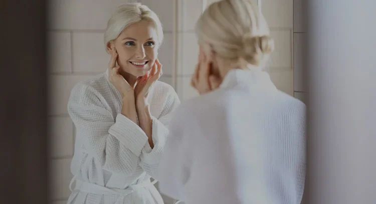 woman in white robe smiling in the mirror at Prime Facial Plastic Surgery