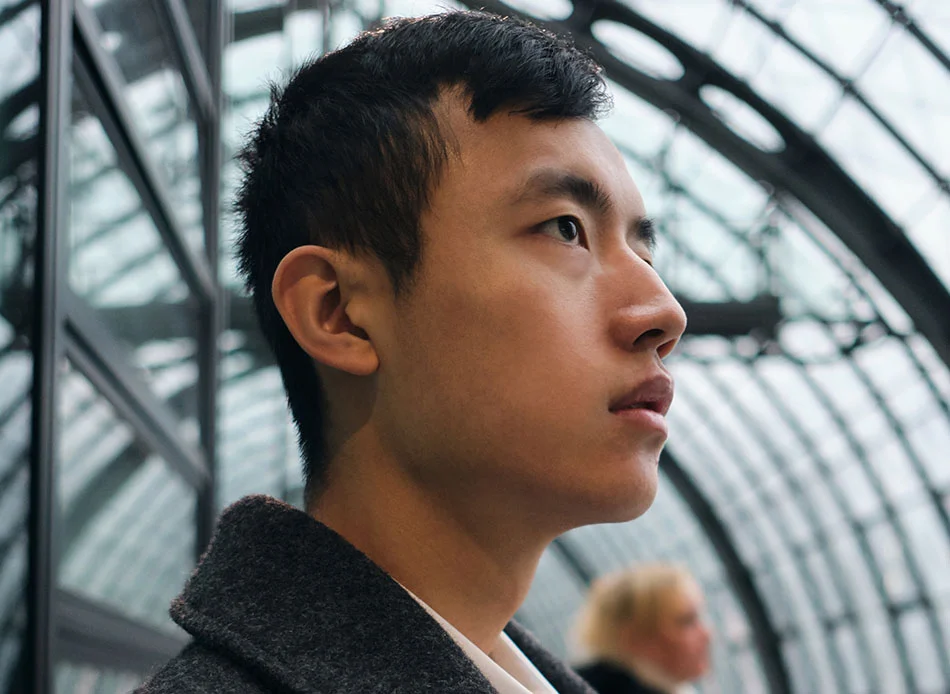 A young Asian man looking thoughtfully into the distance, standing in front of a modern architectural structure - Otoplasty in Bethesda, MD